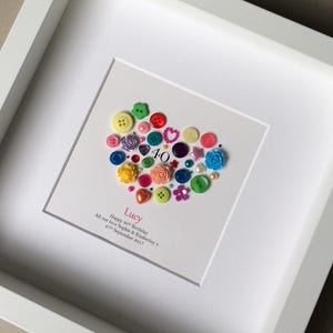 Puede incluir: Un marco blanco con un coraz&oacute;n hecho de botones coloridos. El coraz&oacute;n est&aacute; rodeado de un fondo blanco con el texto "Lucy Happy 40th Birthday All our love Sophie & Kimberley x 27th September 2017".
