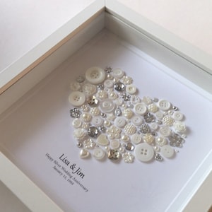 May include: A white frame with a white background and a heart shape made of white and silver buttons. The text "Lisa & Jim Happy Silver Wedding Anniversary January 13, 1998" is written below the heart.
