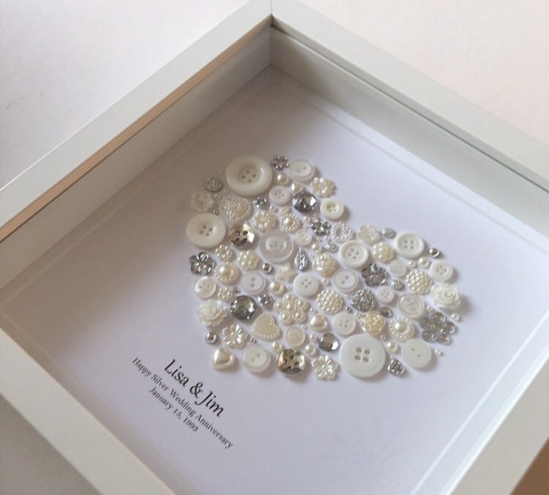 May include: A white frame with a white background and a heart shape made of white and silver buttons. The text "Lisa & Jim Happy Silver Wedding Anniversary January 13, 1998" is written below the heart.