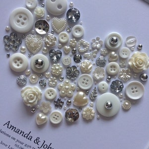 May include: A white card with a heart shape made of white and silver buttons, pearls, and other embellishments. The text "Amanda & John" and "Congratulations on your 25th Wedding Anniversary" is written on the card.
