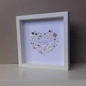 May include: A white frame with a white background and a heart made of white and silver buttons. The text "Congratulations on your 15th Wedding Anniversary Claire & Darren 1st April 2016" is written in silver lettering.