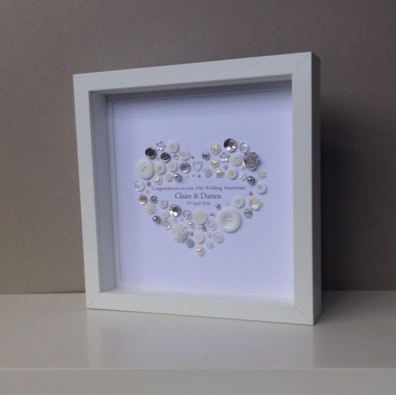 May include: A white frame with a white background and a heart made of white and silver buttons. The text "Congratulations on your 15th Wedding Anniversary Claire & Darren 1st April 2016" is written in silver lettering.