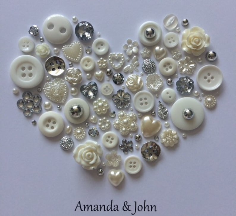 May include: A white heart-shaped arrangement of buttons, pearls, and other embellishments. The heart is surrounded by a white background and the text "Amanda & John" is written below the heart.