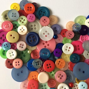 May include: A heart-shaped arrangement of colorful buttons in various sizes and styles. The buttons are made of plastic, metal, and fabric. Some buttons have four holes, while others have two. The buttons are arranged in a random pattern, creating a visually appealing and textured design.
