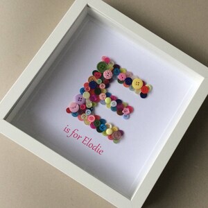May include: A white frame with a white background. The letter "E" is spelled out using colorful buttons. The text "is for Elodie" is written below the letter.