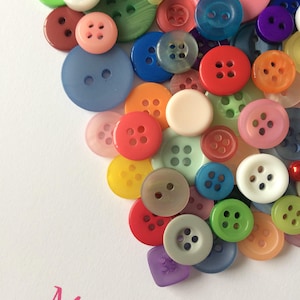 May include: A heart-shaped arrangement of colorful buttons in various sizes and textures. The buttons are arranged in a random pattern, creating a visually appealing and unique design. The text "Miss B" is written in pink lettering at the bottom of the image.