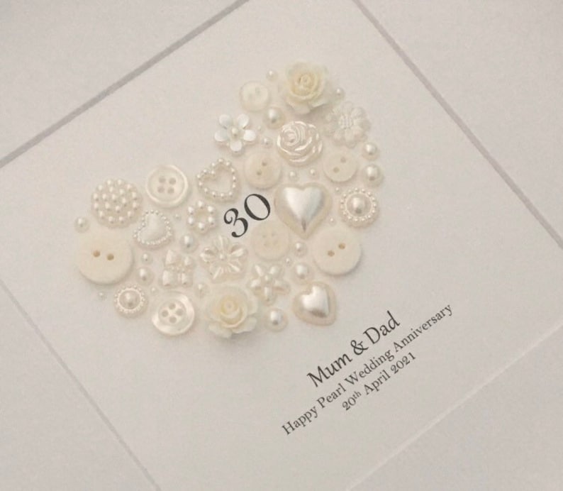 May include: A white card with a heart made of white buttons, pearls, and other embellishments. The number 30 is in the center of the heart. The text "Mum & Dad Happy Pearl Wedding Anniversary 20th April 2021" is below the heart.