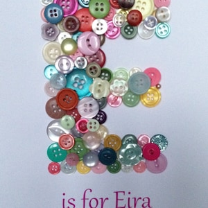 May include: A white card with the letter "E" spelled out using colorful buttons. The text "is for Eira" is written below the letter. The text "Happy 1st Birthday 3rd February 2016" is written below the name.