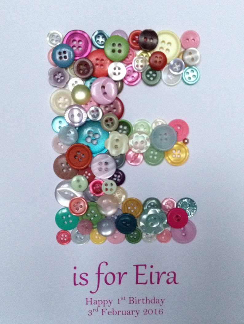 May include: A white card with the letter "E" spelled out using colorful buttons. The text "is for Eira" is written below the letter. The text "Happy 1st Birthday 3rd February 2016" is written below the name.
