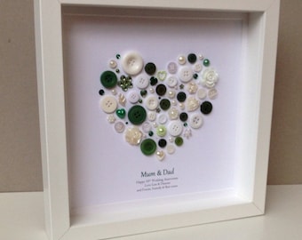 May include: A framed artwork featuring a heart shape composed of various buttons in shades of green, white, and cream. The buttons are of different sizes and textures, creating a textured effect. The frame is white, and the artwork includes text at the bottom.