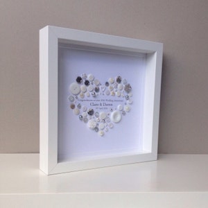 May include: White framed picture with a white background. A heart shape is created with white and silver buttons. The text reads "Congratulations on your 10th Wedding Anniversary Claire & Darren 1st April 2016".