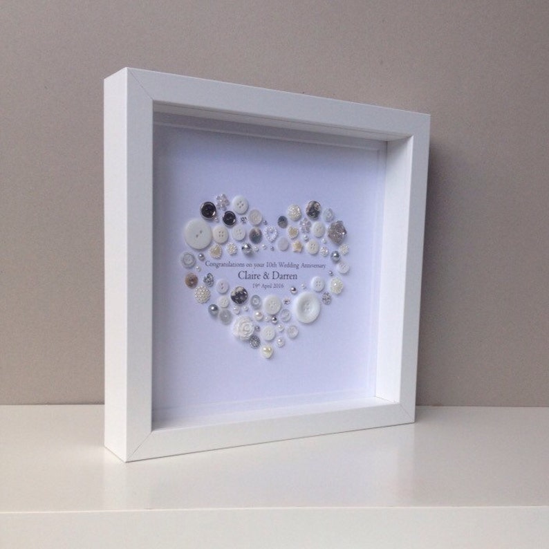 May include: White framed picture with a white background. A heart shape is created with white and silver buttons. The text reads "Congratulations on your 10th Wedding Anniversary Claire & Darren 1st April 2016".
