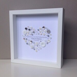 May include: A white frame with a white background and a heart shape made of white and silver buttons. The text "Congratulations on your 10th Wedding Anniversary Claire & Darren 19th April 2016" is written in silver lettering.