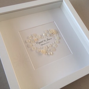 May include: A white frame with a white background and a heart made of white buttons, pearls, and flowers. The text "Crystal & Jason September 2017" is written in black ink below the heart.