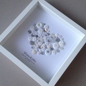 May include: A white frame with a white background. A heart shape is created with a variety of white and silver buttons. The text "Silvana & John Happy Silver Wedding Anniversary June 20, 2014" is written below the heart.