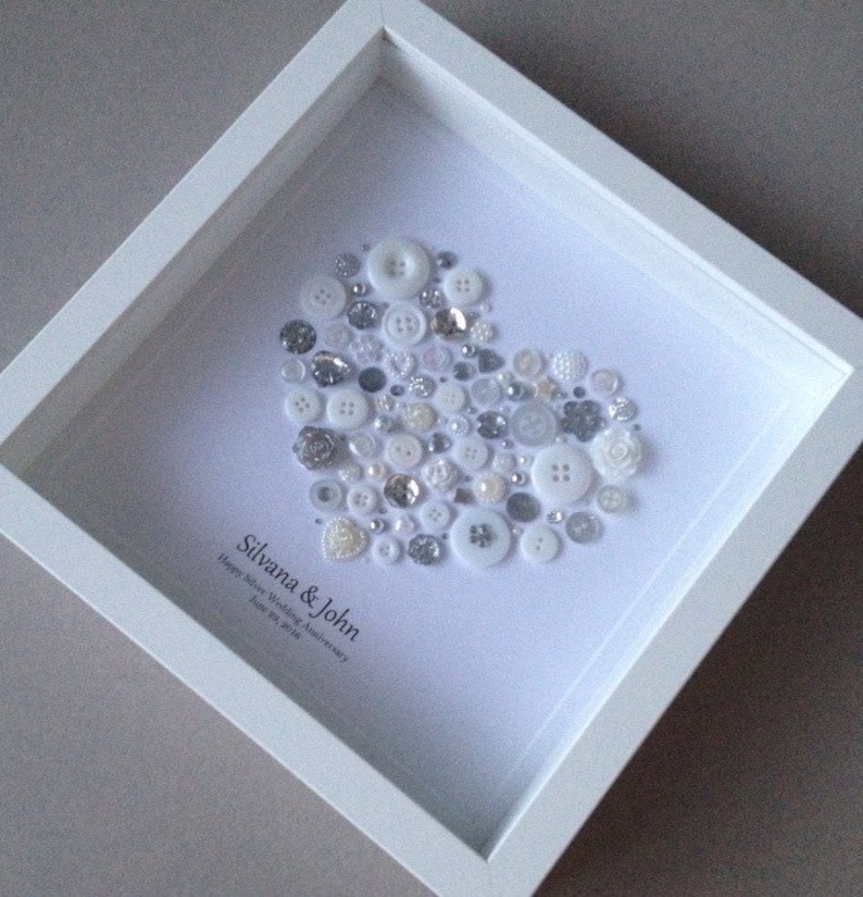 May include: A white frame with a white background. A heart shape is created with a variety of white and silver buttons. The text "Silvana & John Happy Silver Wedding Anniversary June 20, 2014" is written below the heart.