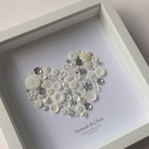May include: White framed picture with a heart made of white and silver buttons, pearls, and beads. The text "Hannah & Chris Happy Wedding Day 03.08.2018" is below the heart.