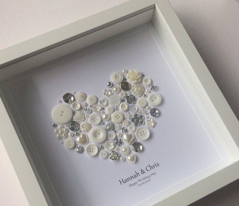May include: White framed picture with a heart made of white and silver buttons, pearls, and beads. The text "Hannah & Chris Happy Wedding Day 03.08.2018" is below the heart.