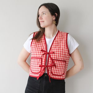 Può includere: Gilet a quadretti rossi e bianchi con finiture rosse e due tasche frontali. Il gilet ha fiocchi rossi per la chiusura. Indossato sopra una maglietta bianca e pantaloni neri.