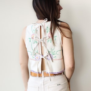Puede incluir: Top blanco sin mangas con un diseño bordado floral y una espalda con cordones. El top es corto y tiene un cinturón de cuero marrón.