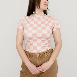 May include: A pink and white checkered top with a mock neck and short sleeves. The top has a ruffled trim around the neckline and sleeves. The model is wearing brown pants.
