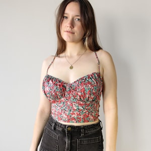 Peut inclure: Une femme portant un haut court à imprimé floral rouge et vert avec de fines bretelles et un décolleté en cœur. Le haut est cintré à la taille et présente un détail plissé au niveau de la poitrine. Elle porte également un pantalon en jean noir.