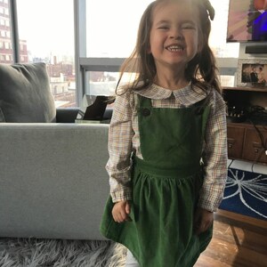 May include: A young girl wearing a green corduroy overall dress with a plaid shirt underneath. The dress has a Peter Pan collar and a button closure. The girl is smiling and has long brown hair.