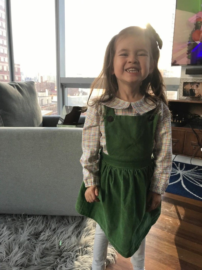 May include: A young girl wearing a green corduroy overall dress with a plaid shirt underneath. The dress has a Peter Pan collar and a button closure. The girl is smiling and has long brown hair.