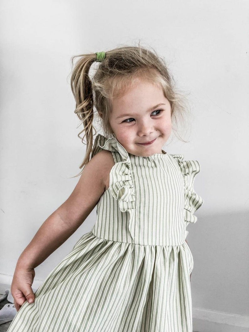 May include: A light green and white striped dress with ruffled sleeves. The dress is sleeveless and has a gathered skirt. The child is wearing a green hair tie.