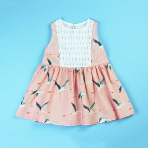 May include: A sleeveless pink dress with a white panel featuring blue raindrop patterns. The dress is adorned with a repeating pattern of flying birds in teal, black, and red. The dress has a gathered skirt and a button closure at the back of the neck.