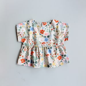 May include: A white cotton blouse with a floral print featuring red, pink, blue, and yellow flowers. The blouse has short sleeves and a ruffled hem.