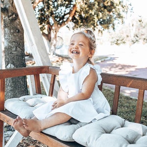May include: A young child wearing a white dress with ruffled sleeves sits on a wooden bench with gray cushions. The child is smiling and laughing, with a background of trees and sunlight.