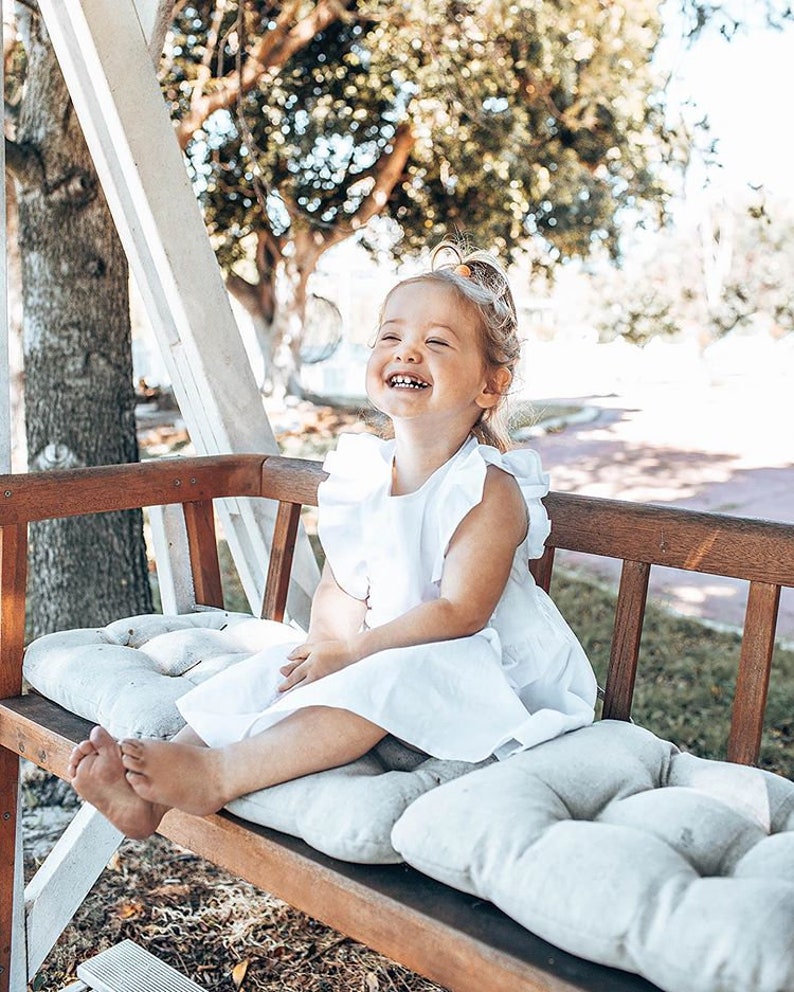 May include: A young child wearing a white dress with ruffled sleeves sits on a wooden bench with gray cushions. The child is smiling and laughing, with a background of trees and sunlight.