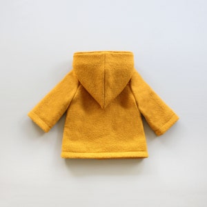 Puede incluir: Una chaqueta amarilla con capucha de textura suave y peluda. La chaqueta tiene un dise&ntilde;o sencillo con capucha y mangas largas.