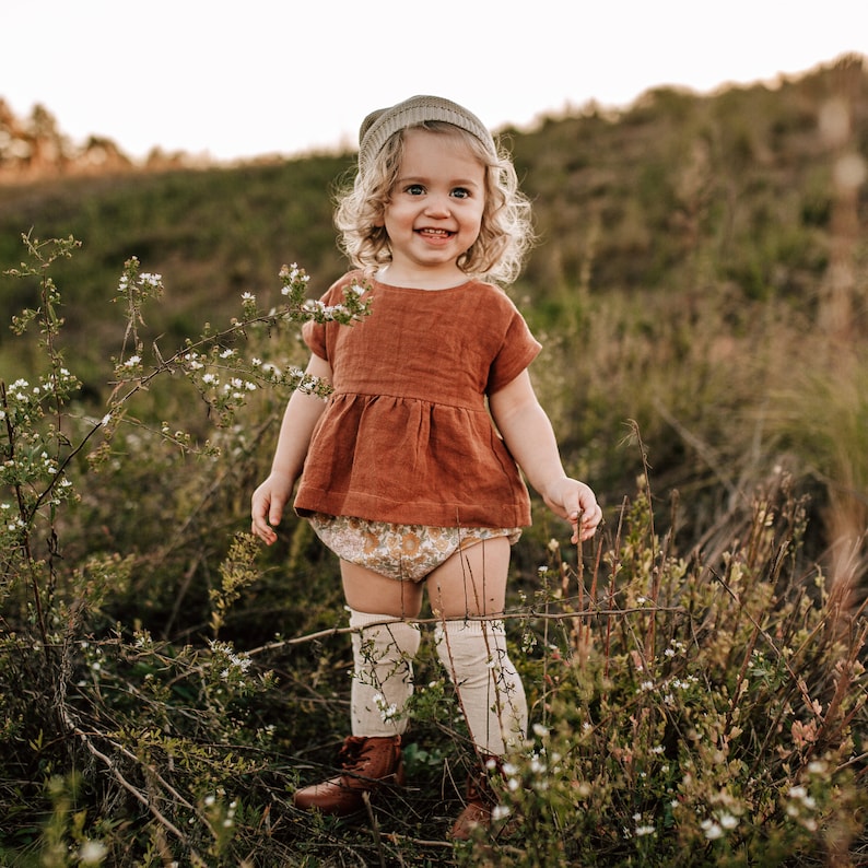 May include: A young child wearing a brown linen dress with a white floral print, a brown beanie, and white knee-high socks. The child is standing in a field of tall grass and wildflowers.