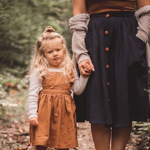 May include: A young girl in a brown dress holds hands with a woman wearing a brown top and a dark blue skirt with brown buttons. They are standing on a path in a forest.