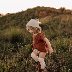 May include: A young child wearing a beige knitted hat with ear flaps, a rust-colored dress, and white knee-high socks walks through a field of tall grass.
