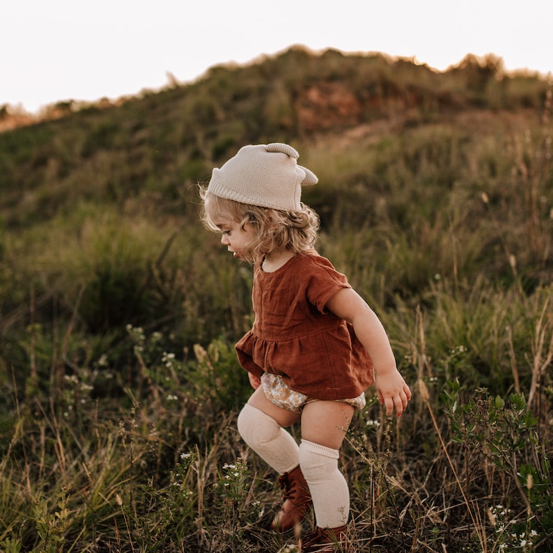 May include: A young child wearing a beige knitted hat with ear flaps, a rust-colored dress, and white knee-high socks walks through a field of tall grass.