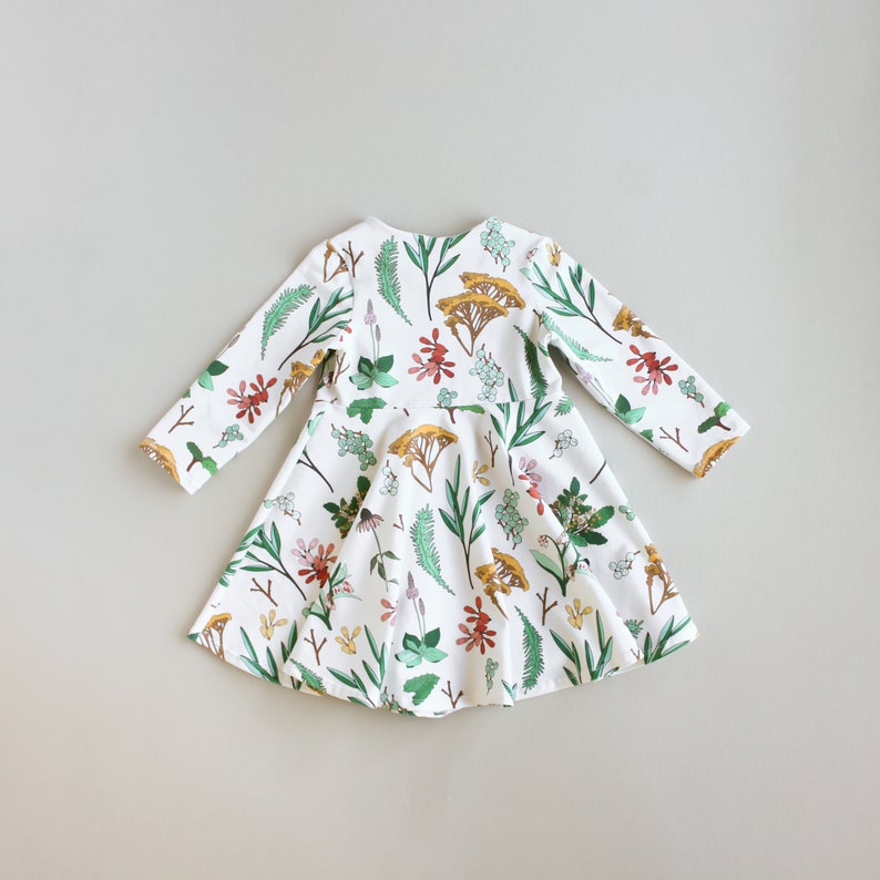 May include: A white dress with long sleeves and a floral print featuring green leaves, red, yellow, and blue flowers. The dress has a flared skirt.