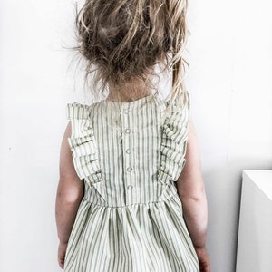 May include: A child wearing a green and white striped dress with ruffled sleeves and a button-down back. The child's hair is pulled back with a green hair tie. The dress has a flared skirt.