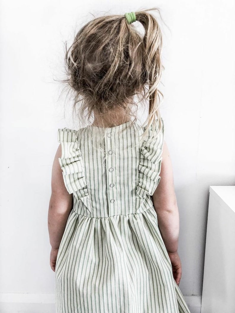 May include: A child wearing a green and white striped dress with ruffled sleeves and a button-down back. The child's hair is pulled back with a green hair tie. The dress has a flared skirt.