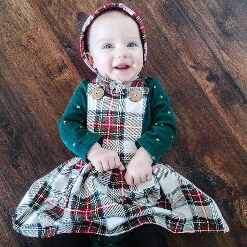 May include: A baby wearing a green and white plaid dress with a matching green hooded hat. The dress has a white background with red and green plaid accents. The baby is smiling and looking at the camera.