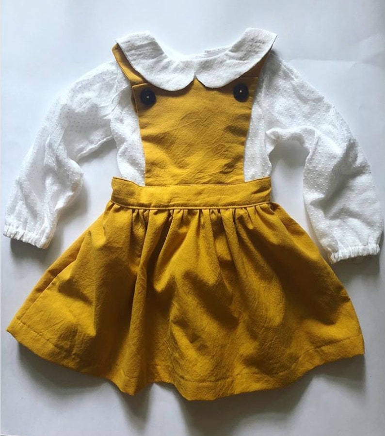May include: A yellow overall dress with a white collared shirt underneath. The dress has two black buttons on the front and a gathered skirt.