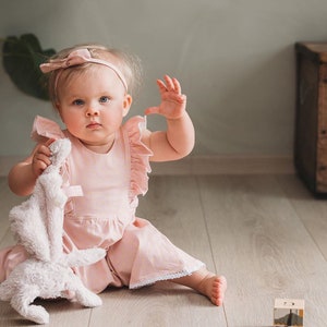 May include: A baby wearing a light pink romper with ruffle sleeves and a matching headband. The baby is holding a white stuffed bunny and sitting on a wooden floor. A wooden block is in the foreground.