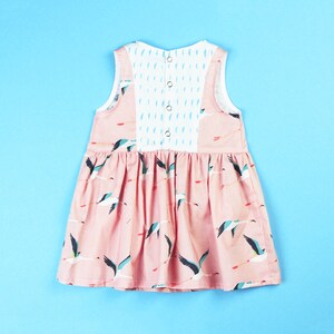 May include: A sleeveless pink dress with a pattern of flying birds. The back of the dress features a white panel with blue accents and three buttons. The dress is designed for children.