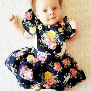 May include: A baby girl wearing a navy blue floral dress with a white bodice and ruffle sleeves. She is wearing a light blue bow in her hair.