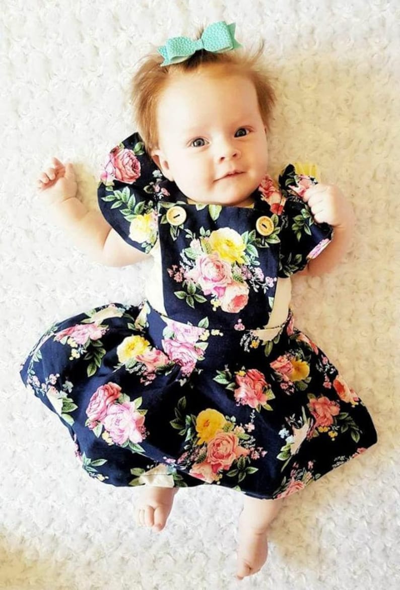 May include: A baby girl wearing a navy blue floral dress with a white bodice and ruffle sleeves. She is wearing a light blue bow in her hair.