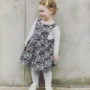 May include: A young girl wearing a white long-sleeved shirt and a black and white floral overall dress. She is wearing white tights and white sneakers. She has a yellow hair clip in her hair.