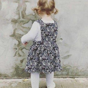 May include: A young child wearing a white long-sleeved shirt and a dark blue and white floral print overall dress. The child has their hair pulled back in pigtails and is standing in front of a textured wall.