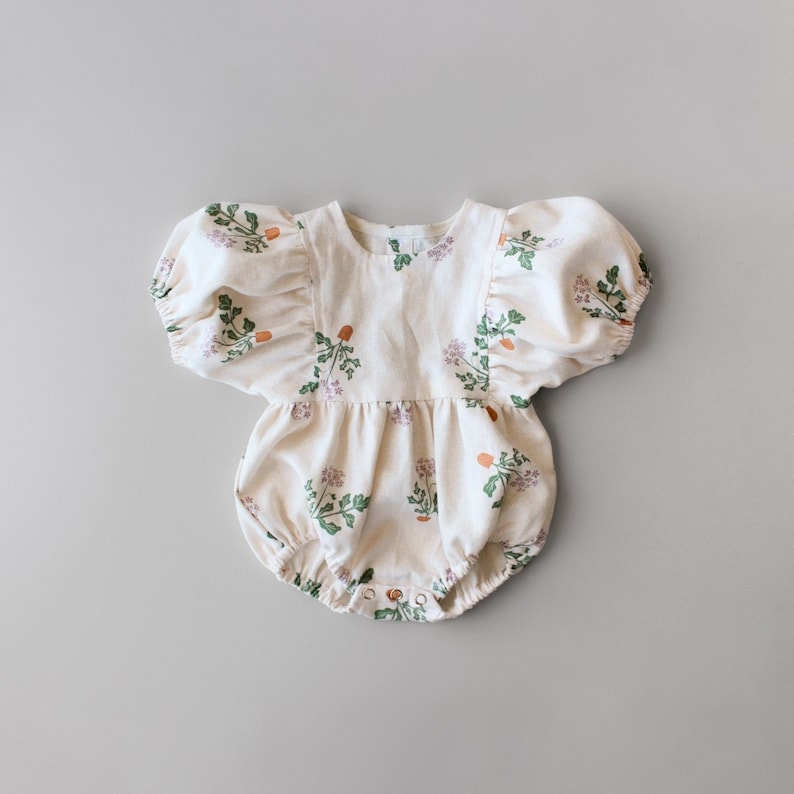 May include: A cream-colored baby onesie with a floral print featuring green leaves, pink flowers, and orange berries. The onesie has puffed sleeves and a ruffled bottom.